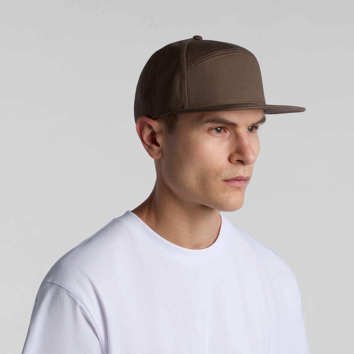 A person wearing a brown flat-brimmed cap and a white t-shirt stands against a plain gray background, looking to the side.