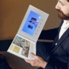 A person with a beard is holding an open brochure featuring a small screen playing a video. The brochure displays images and text about interior design. The person is wearing a dark jacket and sitting on a light-colored surface.