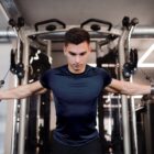 A man wearing an Energetic Tee is working out at the gym on a cable machine, concentrating as he pulls the cables with both arms extended. Gym equipment and lights in the background emphasize his dedication.
