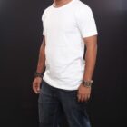 A man wearing an Emblem Tee and dark jeans stands against a black background. With a short beard, he sports a watch and bracelet on his wrists.