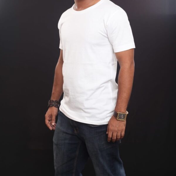 A man wearing an Emblem Tee and dark jeans stands against a black background. With a short beard, he sports a watch and bracelet on his wrists.