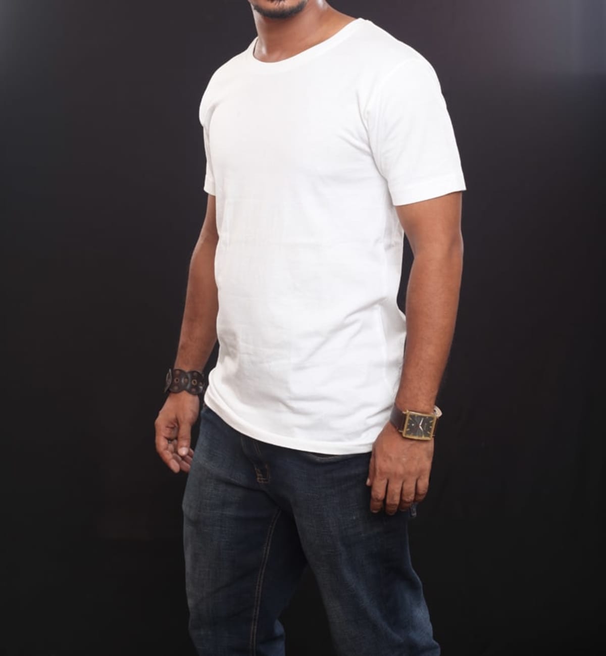 A man wearing an Emblem Tee and dark jeans stands against a black background. With a short beard, he sports a watch and bracelet on his wrists.