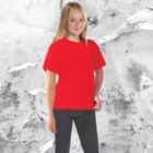A young girl with long blonde hair stands smiling, wearing a bright red Core Junior Tee and dark pants, set against an abstract grey and white backdrop.