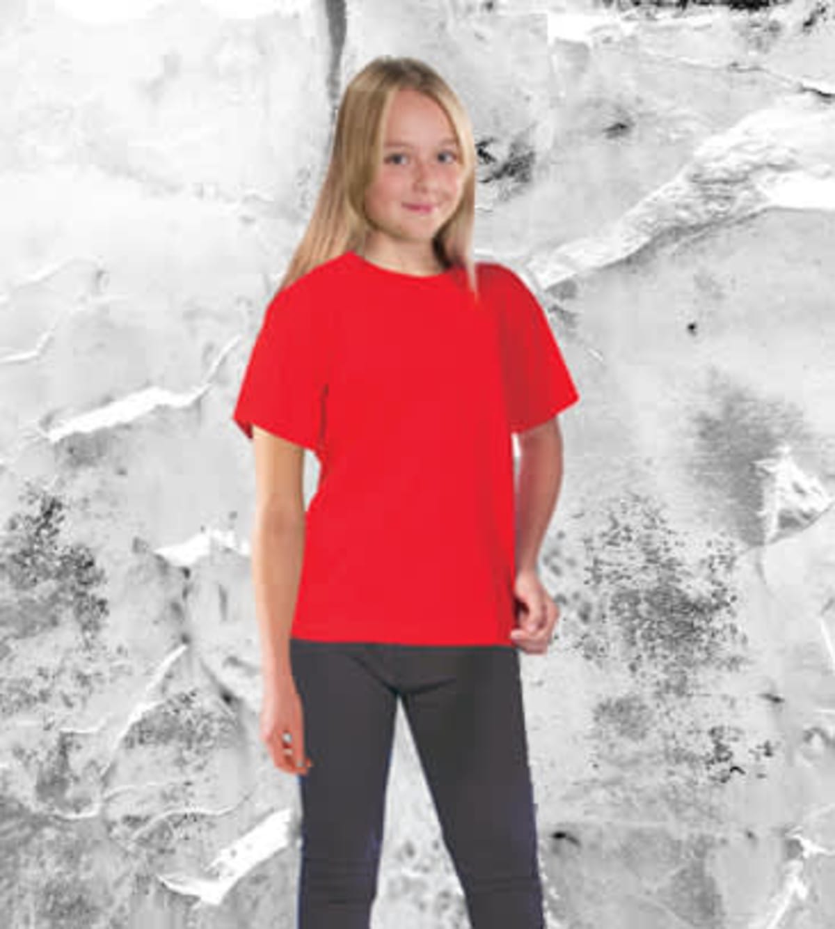 A young girl with long blonde hair stands smiling, wearing a bright red Core Junior Tee and dark pants, set against an abstract grey and white backdrop.