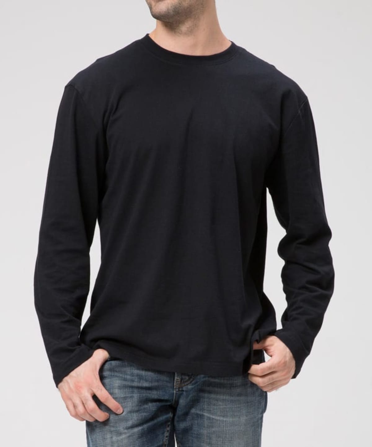 Wearing a Signature L/S Tee in black and blue jeans against a light gray background, they stand with one hand in their pocket, head slightly turned to obscure their face.