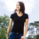 A woman wearing the Ignite Tee and jeans smiles outdoors, framed by trees and a blue sky with clouds.