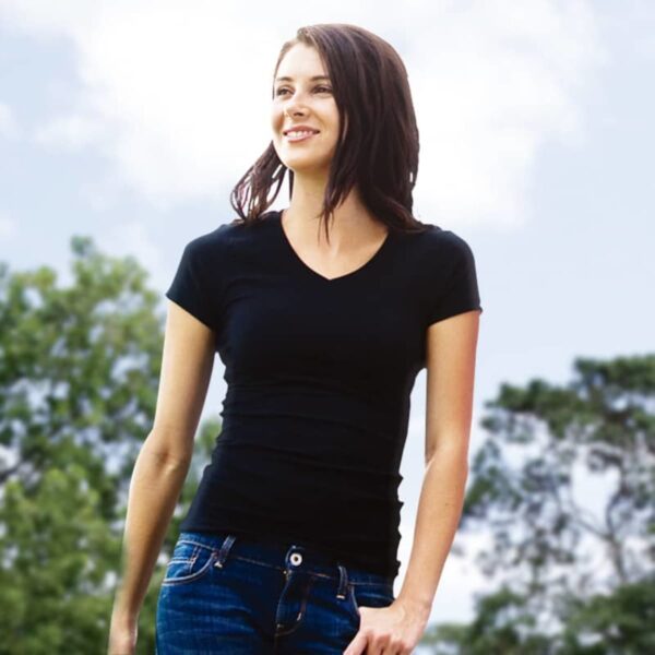 A woman wearing the Ignite Tee and jeans smiles outdoors, framed by trees and a blue sky with clouds.
