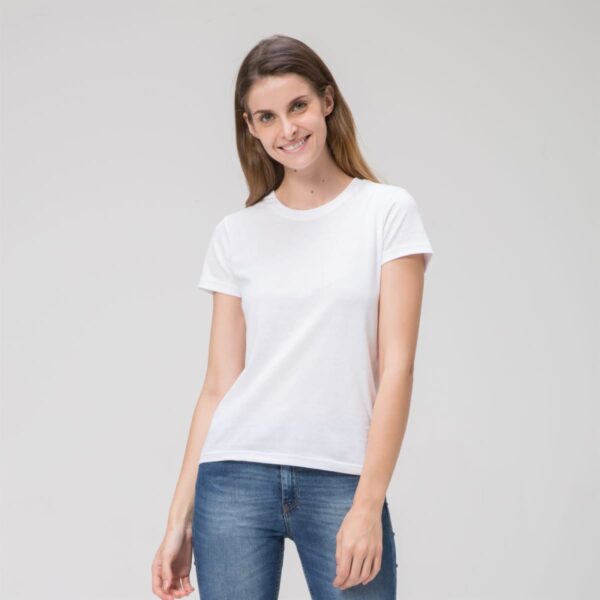 A person smiles against a plain backdrop in a Prime Ladies Tee and blue jeans, their long hair cascading naturally, enhancing their relaxed pose with one hand slightly bent.