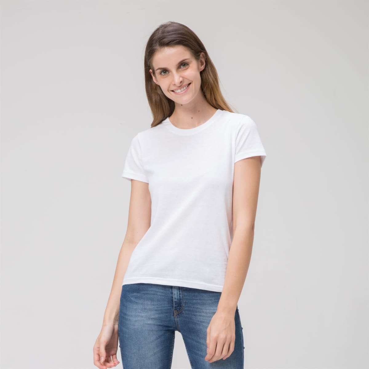 A person smiles against a plain backdrop in a Prime Ladies Tee and blue jeans, their long hair cascading naturally, enhancing their relaxed pose with one hand slightly bent.