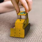 A persons hand is holding a small toy bulldozer with the word FORCE and a logo on the top. The toy is yellow with black tracks, and it is placed on a carpeted surface.