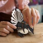 A person assembles a colorful paper bird model on a wooden table. Their hands delicately hold the wings, which are painted in black and white patterns. The person wears a pink top with ruffled sleeves.