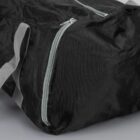 A black duffel bag with gray handles and a light blue zipper is shown. The bag is crumpled slightly, indicating its made of a soft, flexible material. Its set on a gray surface.