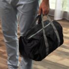 A person in light gray pants holds a black duffel bag by its gray handles. The background shows a wooden floor and a sheer curtain.