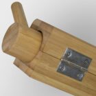 Close-up of a wooden object featuring a smooth cylindrical handle inserted into a rectangular wooden base with a metal hinge. The wood has a natural finish, showcasing its grain pattern. The background is plain and light-colored.