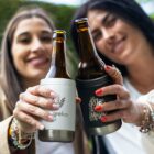 Two smiling people raise beer bottles with patterned cozies for a toast. The focus is on the bottles in the foreground, while the background is slightly blurred. The scene suggests a casual, outdoor celebration.