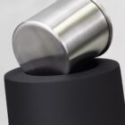 A stainless steel cylinder resting at an angle on top of a cylindrical black stand with a matte finish, against a plain gray background.