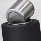 A shiny metal cup is tilted at an angle, resting against the opening of a matte black cylindrical container. The image focuses on the contrast between the metallic and matte textures.