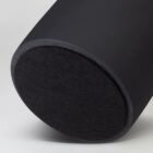 Close-up of a black cylindrical speaker with a rounded bottom resting on a light gray surface. The speakers fabric texture and sleek design are clearly visible.