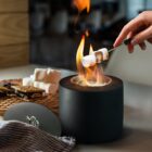 A hand holds a skewer with a marshmallow over a small tabletop fire for roasting. Nearby are graham crackers, chocolate, and a stack of marshmallows, set on a cooling rack, suggesting smores preparation. A striped cloth lies on the counter.