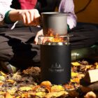 A person sitting on a bed of autumn leaves is using a small camping stove with a visible flame to heat a pot. The stove has The Forest Adventurer logo on it.