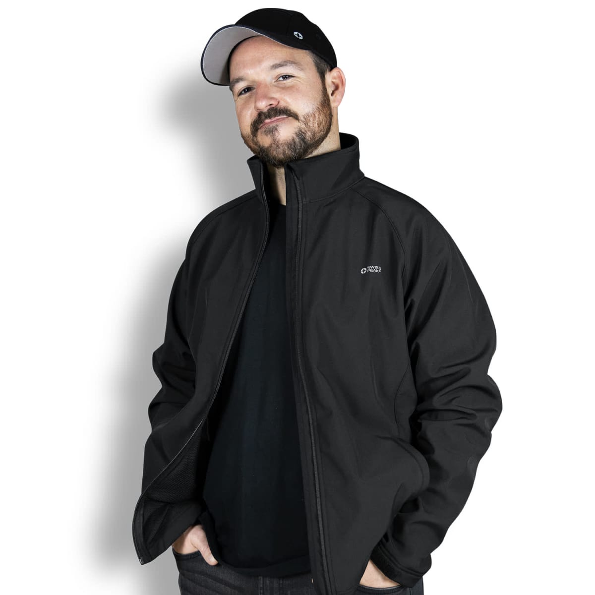 A person wearing a black jacket and black cap stands against a white background, smiling with hands in pockets. The jacket has a small logo on the left chest.