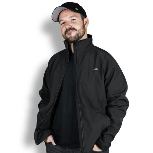 A bearded man in a black baseball cap and jacket stands against a white background. He has a slight smile and his hands are in his pockets.