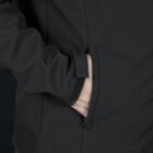 A person wearing a black jacket with their hand in the pocket. The jacket features a zippered pocket and a velcro strap on the cuff. The background is dark, highlighting the details of the jackets fabric and design.