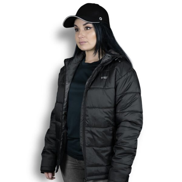 A person wearing a black puffer jacket and a black cap stands against a white background. They have long dark hair and are looking off-camera.