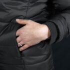 A person wearing a black puffer jacket with their left hand in the pocket. They are wearing a ring on their ring finger. The focus is on the hand and jacket texture.