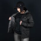 A person wearing a black cap and a dark puffer jacket is looking down and opening the jacket slightly to reveal the inside lining. They are standing against a dark background.
