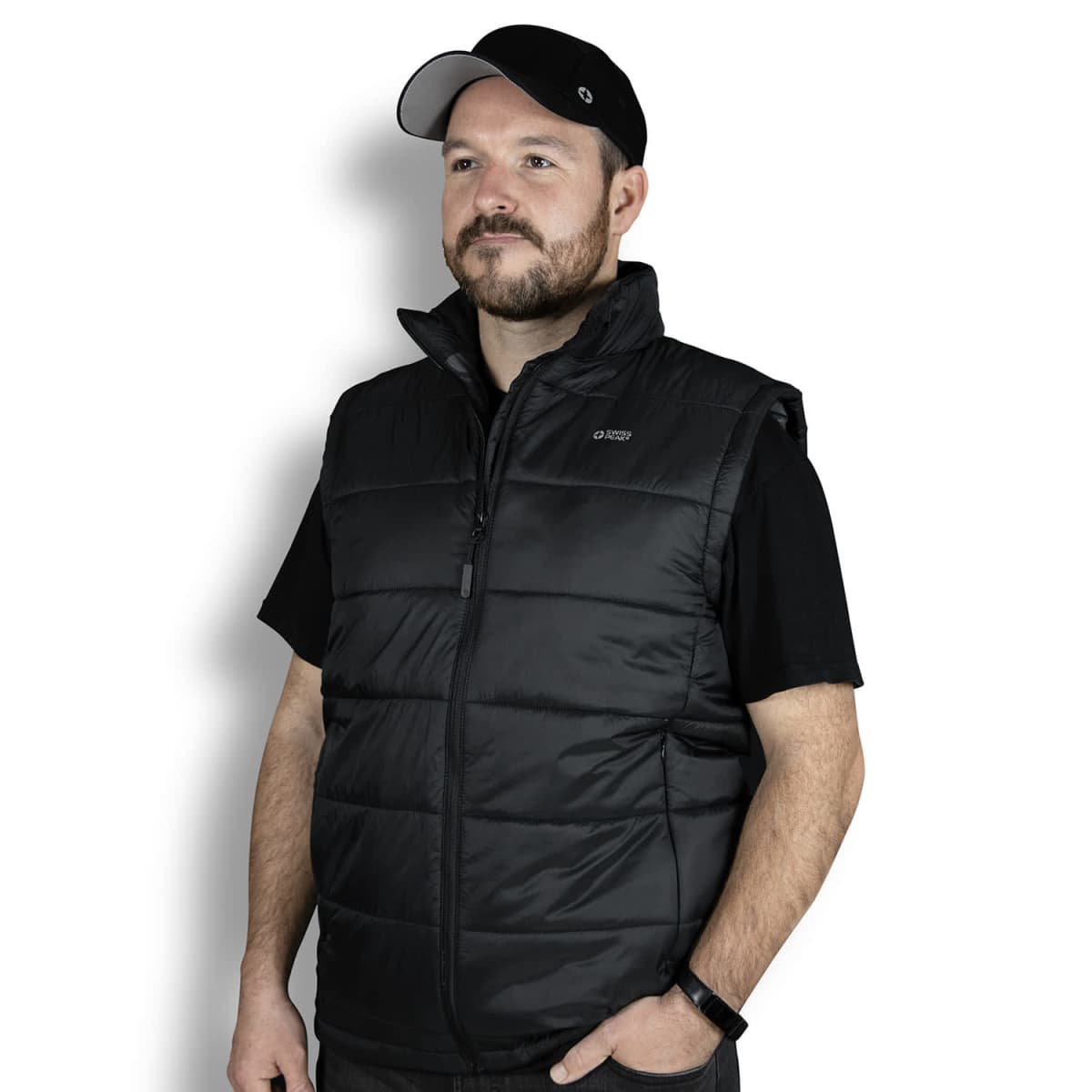 A man wearing a black cap, black T-shirt, and black puffer vest stands against a white background. He has a beard and is looking to the side. His left hand is in his pocket.