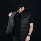 A person wearing a black cap and a sleeveless puffer jacket over a black shirt is looking down while holding open the jacket to show its patterned interior lining. The background is solid black.