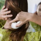 A person brushing their long brown hair while looking in a mirror. They are wearing a green patterned shirt. The background is blurred, focusing on the hand holding a white hairbrush.