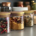 Three glass jars with wooden lids are filled with colorful spices, including peppercorns and star anise. The labels read Urban Finds Market. The jars are arranged in a row on a gray countertop, with more jars in the background.