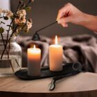 A hand using a candle snuffer to extinguish a lit candle on a wooden table. Two candles sit in a black tray beside a pair of wick trimmers. In the background, there are flowers in a vase and a blurred-out room setting.