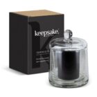 A black candle in a clear glass holder with a lid, placed next to a black box labeled keepsake collection. The box has a brown top and text detailing the products features.