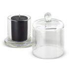 A black candle in a clear glass holder is placed next to an empty glass dome with a knob handle, both sitting on a white surface. The dome is open, positioned slightly to the right of the candle.