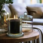A lit candle in a glass holder sits on a wooden table next to an empty glass cloche. In the background, there is a blurred view of a sofa with cushions, creating a cozy atmosphere.
