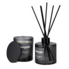 A black candle jar labeled Whiskey + Orange sits next to a black reed diffuser with the same label. The diffuser has several black reeds fanned out and a loose black lid is beside the candle. Both are branded keepsake.