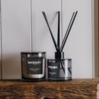 A candle and a reed diffuser labeled Keepsake Whiskey + Orange are displayed on a wooden shelf against a white paneled background.