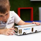 A young boy intently plays with a white toy bus labeled People Travel. The background is slightly blurred with hints of colorful furniture. The boy is wearing a white and gray striped shirt.