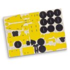Image of a punch-out board with various cardboard pieces, including yellow shapes and black circular tokens. The pieces appear to be part of a board game or model kit, arranged in a grid-like pattern on the sheet.