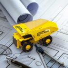 A miniature yellow dump truck labeled Rock Solid is placed on top of architectural blueprints, surrounded by drafting tools including a compass, ruler, and pencil.