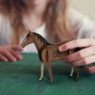 A child holds a toy horse with Wild West Rodeo printed on its side. The horse is made of wood, and the child is seated at a green table. The focus is on the horse.