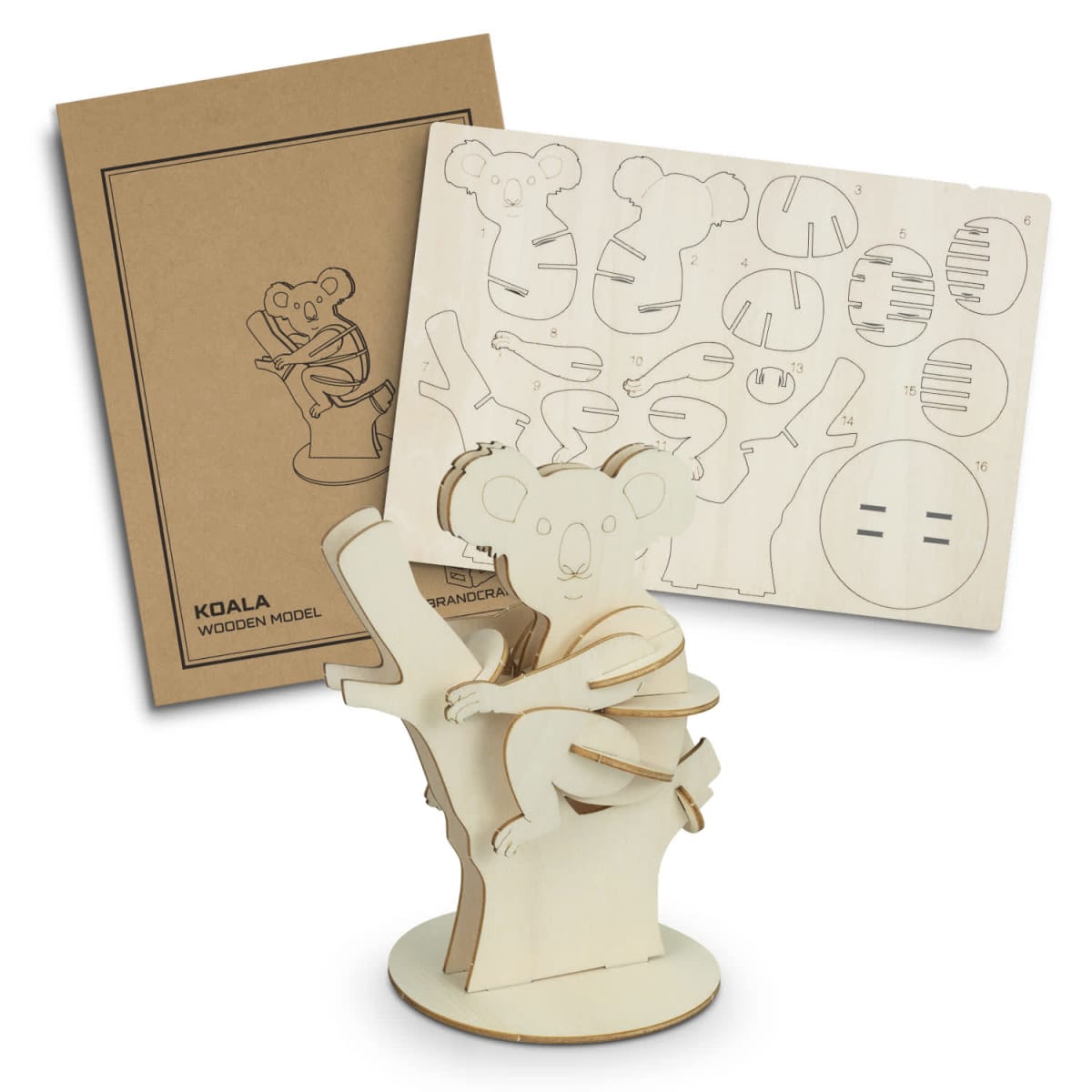 Wooden koala model kit with a completed koala on a tree stump and an unassembled wooden board visible in the background. The kit includes instructions and cut-out pieces for assembly.