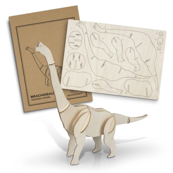 A wooden Brachiosaurus model stands assembled in front of its disassembled kit. The kit includes a cardboard cover with a dinosaur illustration and a wooden sheet showing cut-out puzzle pieces.