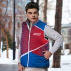 A man wearing a red and blue sports vest stands outdoors. The background features a snowy scene with trees and buildings.