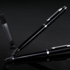 A sleek black pen with silver accents is positioned diagonally against a dark reflective surface, with its cap open at one end. A transparent pen case stands to the left, enhancing the elegant presentation.