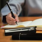 A person writing in a notebook on a wooden desk, holding a black pen. Two black pen boxes are nearby, one open with a pen and the other closed. The person wears a light-colored sleeve.