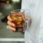 A person in a white shirt holding a glass of amber liquid with ice cubes. The background is out of focus, highlighting the drink in the foreground.